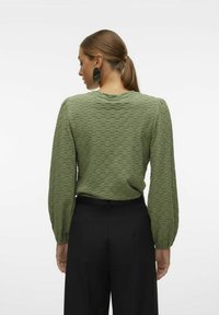 Top verde testurizzato con lunghe maniche a palloncino, scollo rotondo e un motivo sottile. Abbinato a pantaloni neri a vita alta per un look contrastante.