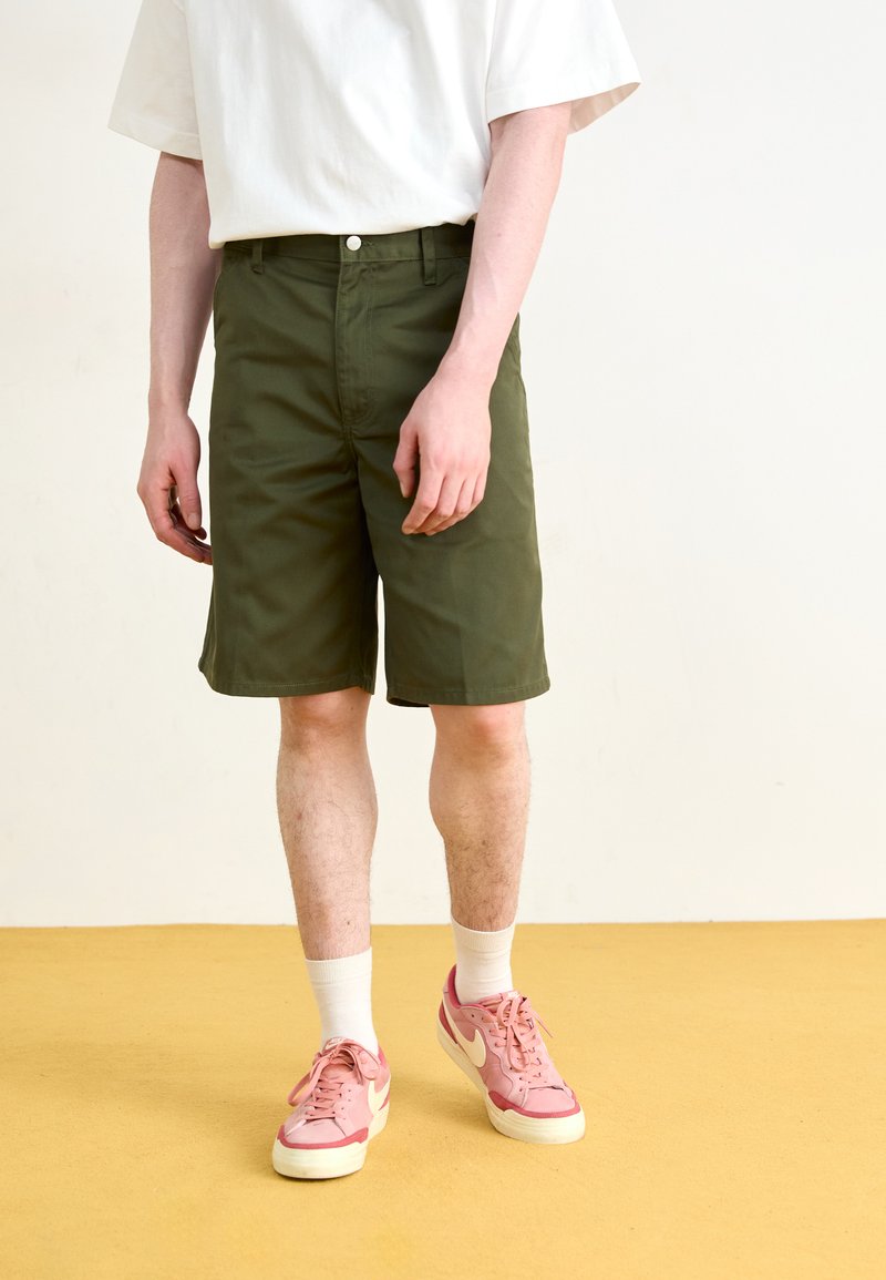 Pantaloni scurți din bumbac verde cu o croială lejeră, închidere cu nasture și buzunare laterale, asortați cu adidași roz și șosete albe până la gleznă.