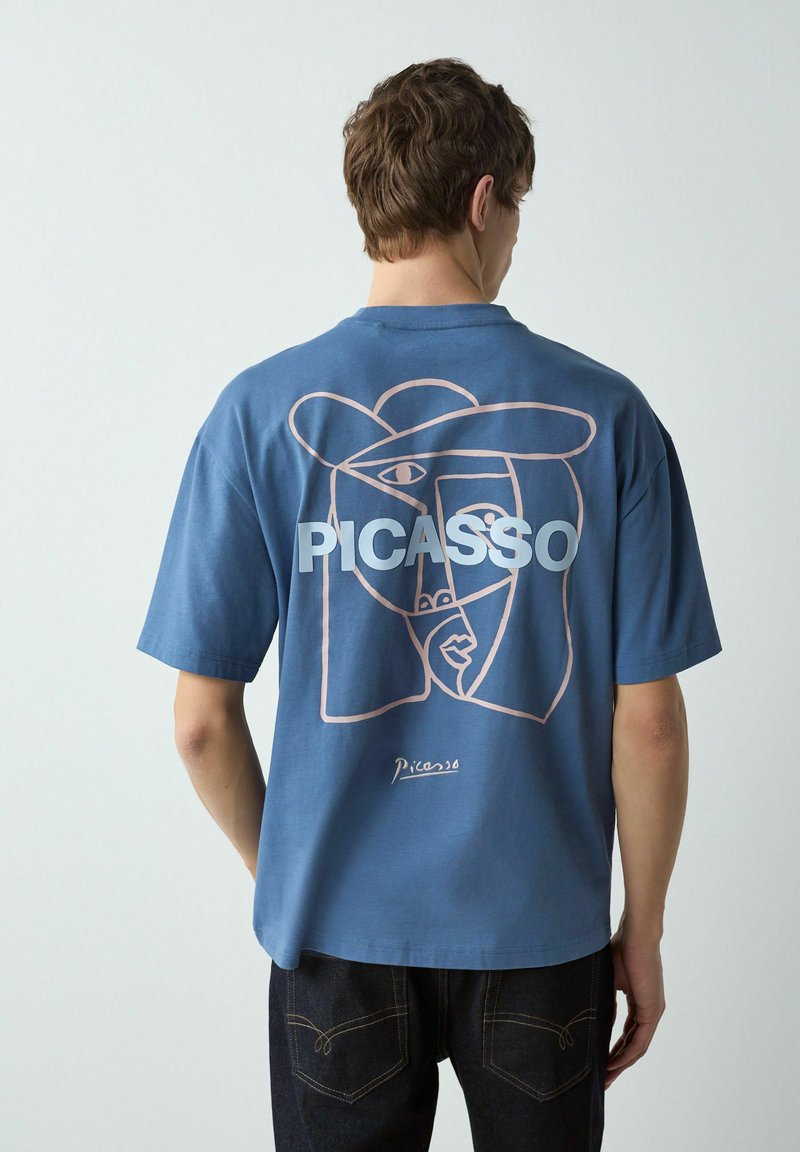 Mand vendt væk iført en blå T-shirt med en abstrakt stregtegning af et ansigt og ordet "PICASSO" på ryggen.