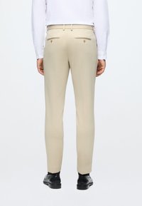 Smėliniai pritaikyti kelnės su dviem užpakaliniais kišenėmis ir auksiniais mygtukais, derinami su juodais batais ir balta ilgomis rankovėmis marškiniais. Sklandus tekstūra.