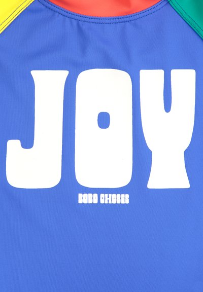 Blått tyg med stora vita bokstäver "JOY" och "BOBO CHOSES" nedan. Utsmyckat med röda, gula och gröna detaljer på axlarna.