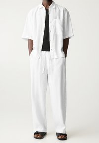 Homme portant un pantalon en lin blanc ample et une chemise à manches courtes assortie sur un t-shirt noir, assortis à des sandales noires, debout les mains dans les poches.