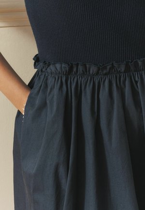 Hand einer Frau in der seitlichen Tasche eines gerafften Marineblauen Rocks, getragen mit einem schwarzen, gerippten Oberteil.