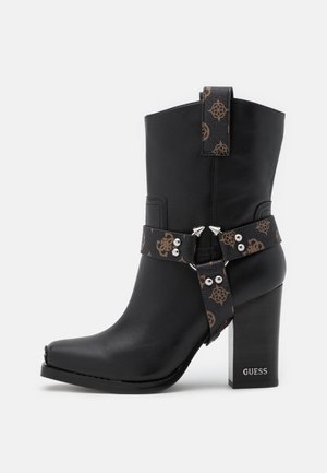 Botte noire en cuir mi-mollet avec talon bloc, décorée de brides marron à motifs avec des clous argentés et le logo GUESS sur le talon.