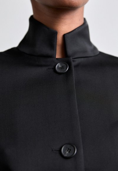 Black tailored jacket featuring a high collar, smooth texture, and two prominent black buttons at the front.