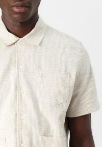 Camisa de manga corta de botones en tejido texturizado beige claro, con un bolsillo en el pecho y un diseño de cuello clásico.