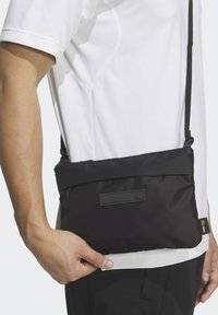 Borsa a tracolla nera con texture liscia in nylon, dotata di una tasca frontale con zip e una patch con il marchio. Tracolla regolabile per maggiore comodità.