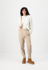 Hvit genser med gule og grønne broderte mønstre, paret med beige bukser og brune semskede ankelstøvler, som står mot en nøytral bakgrunn.