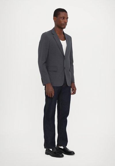 Grauer maßgeschneiderter Blazer mit Revers, zwei Knöpfen und Fronttaschen, kombiniert mit dunklen Jeans und schwarzen Mokassins. Glatte Stoffstruktur.