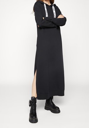 Mujer con vestido largo negro con capucha y abertura lateral, y botas negras de combate con plataforma, de pie con los brazos cruzados.