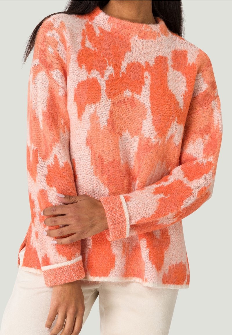Pull en maille douce avec motif abstrait orange et rose clair, manches longues, col rond, porté avec un pantalon de couleur claire.