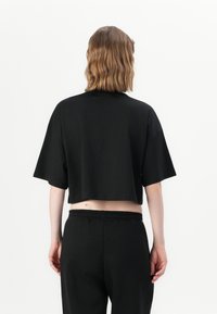 Osoba s ramennou dĺžkou hnedých vlasov, oblečená v voľnom čiernom cropped tričku a čiernych nohaviciach, z pohľadu zozadu na bielom pozadí.