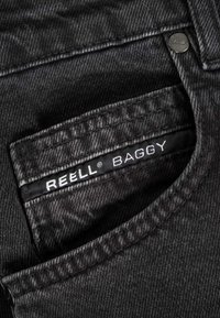 Primer plano del bolsillo de unos vaqueros negros de mezclilla con la etiqueta "REELL BAGGY" y un remache metálico en la esquina superior.