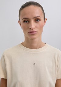 Camiseta de algodón beige con escote redondo, mangas cortas y un pequeño logo bordado en el pecho. Textura suave, corte casual.