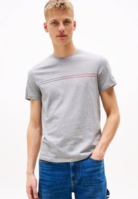 Pelēks kokvilnas t-krekls ar apaļu kakla izgriezumu, īsām piedurknēm un horizontālām sarkanām un zilām svītrām, uz krūškurvja ir teksts "TOMMY JEANS".