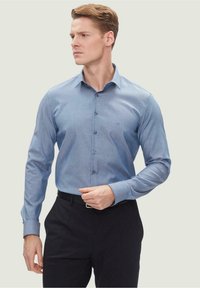 Camicia azzurro chiaro con sottile trama, maniche con bottone e colletto classico, abbinata a pantaloni scuri. Design semplice e sartoriale.