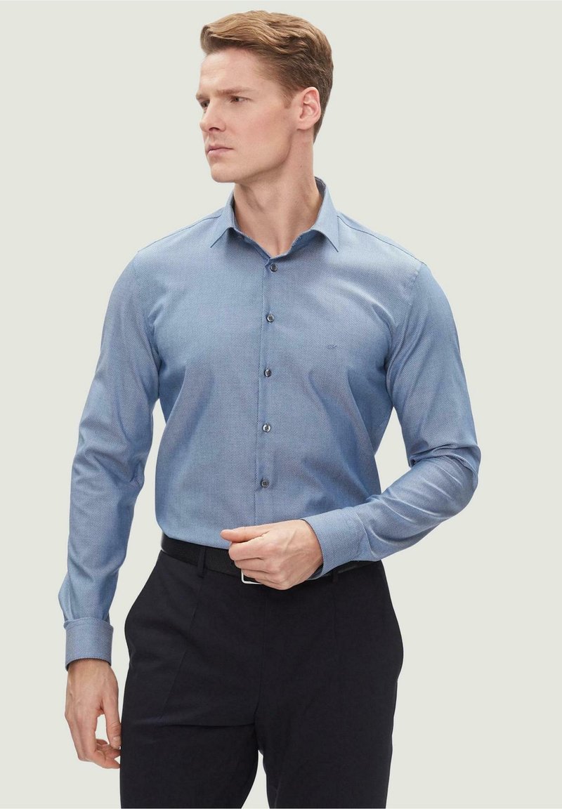 Camicia azzurro chiaro con sottile trama, maniche con bottone e colletto classico, abbinata a pantaloni scuri. Design semplice e sartoriale.