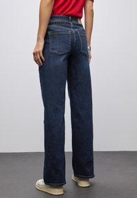 Persona con pantalones vaqueros anchos de color azul oscuro, camisa roja, zapatillas blancas y una pulsera de plata, de pie sobre un suelo gris frente a una pared simple.