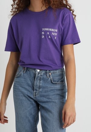 Osoba ubrana w fioletowy t-shirt z napisem „TOMORROW IS A NEW DAY” oraz niebieskie jeansy z wysokim stanem na jednolitym tle.