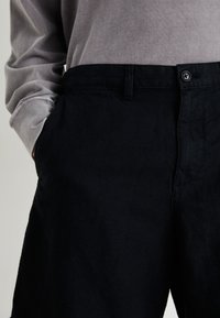 Schwarze Leinenhose mit geradem Schnitt, ausgestattet mit einem einzelnen Knopfverschluss und seitlichen Taschen, kombiniert mit einem hellgrauen Langarmhemd.