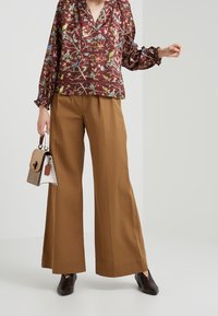 Blouse florale bordeaux avec des poignets élastiques, associée à un pantalon marron ample. Les accessoires comprennent un sac à main structuré marron avec une base blanche.