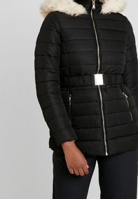 Černá péřová bunda s kapucí lemovanou umělým kožichem, kovovým zipem, páskem v pase a prošívanou strukturou. Má boční kapsy na zip.