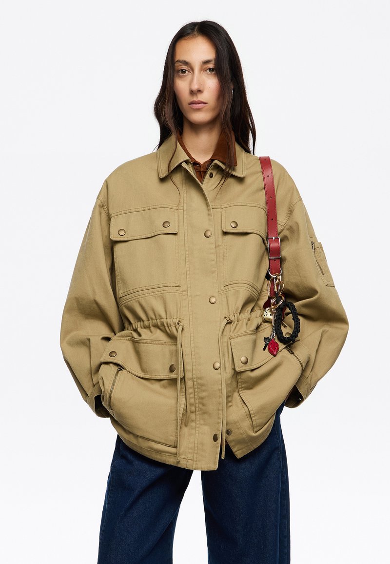 Chaqueta oversized color caqui con cintura ajustable, dos bolsillos en el pecho y cuello. Llevada con una blusa marrón y jeans oscuros, acentuada con accesorios.