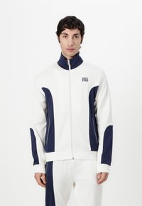Veste blanche zippée avec des accents bleu marine, dotée d'un col montant, de poches latérales et d'un tissu texturé. Logo brodé sur la poitrine.