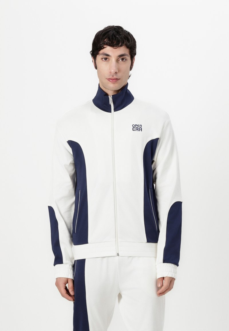 Veste blanche zippée avec des accents bleu marine, dotée d'un col montant, de poches latérales et d'un tissu texturé. Logo brodé sur la poitrine.