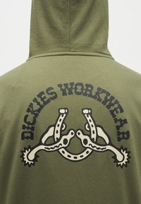 Φούτερ με κουκούλα σε χρώμα ελαιόπρασινο, πίσω όψη με μαύρο κείμενο "Dickies Workwear" πάνω από γραφικό με δύο συνδεδεμένους αγκίδες παπουτσιών.