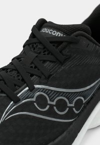 Gros plan sur une chaussure de course Saucony noire avec une tige en mesh, des lacets noirs, un logo argenté et une semelle blanche visible.