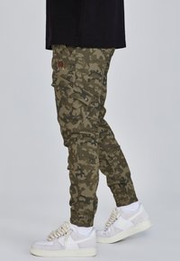 Nohavice typu cargo v zelenej a čiernej maskáčovej vzore, vybavené viacerými vreckami a elastickými manžetami, doplnené o biele tenisky.