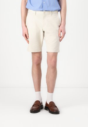 Beige chinos shorts, cotton material, straight cut, front pockets, worn with brown loafers and white socks, displayed on a standing model.