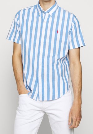 Homme portant une chemise boutonnée à manches courtes, à rayures verticales bleues et blanches, avec un pantalon blanc, une main dans la poche, sur fond clair.