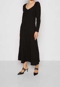 Svart långärmad klänning med texturerad tyg, rundad halsringning och en svängd silhuett. Kombinerad med svarta spetsiga klackskor med runda detaljer.