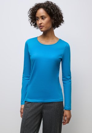Langärmliges, rundhalsiges blaues Oberteil aus glattem Stoff, kombiniert mit strukturierten grauen Hosen. Einfaches Design, keine Muster oder Verzierungen.