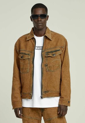 Brown denim jacket with two chest pockets, zip closure, and frayed edges, layered over a white t-shirt; paired with brown pants and sunglasses.