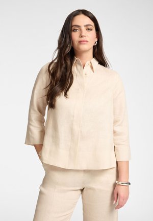 Vrouw met lang donker haar die een beige shirt met driekwart mouwen en bijpassende broek draagt, staand met één hand in de zak tegen een witte achtergrond.