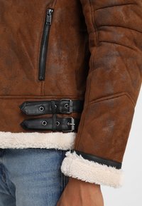 Brun faux suede jakke med et shearling-for, der har sorte læder spænder, en lynlåslomme og syninger langs ærmerne.