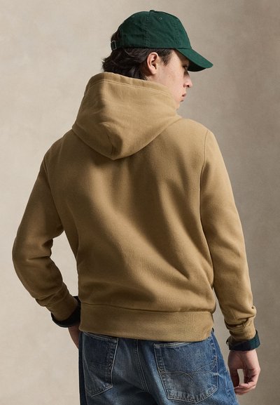 Muž v béžovej mikine s kapucňou, zelenej šiltovke a modrých džínsoch stojí chrbtom k nám a mierne obrátenou hlavou do strany.