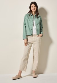 Mentengroene button-up jas, witte t-shirt, beige wijde broek en tan klompen. Gladde stof, comfortabele pasvorm, zichtbare stiksels en zijzakken.