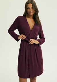 Burgundowa sukienka kopertowa z długimi rękawami, dekoltem w serek i wiązanym w pasie. Wykonana z miękkiego materiału, o gładkiej strukturze i długości do kolan.