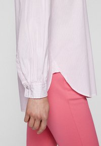 Chemise à rayures rose clair avec un ourlet arrondi et un poignet boutonné, associée à un pantalon côtelé rose vif ajusté. Gros plan sur la vue de côté.