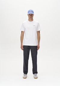 T-shirt bianca a maniche corte con un piccolo logo nero, abbinata a pantaloni grigio scuro e sneakers grigie. Un cappellino blu completa l'outfit.