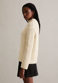 Cremefarbener Strickpullover mit strukturiertem Muster, rundem Halsausschnitt und langen Ärmeln, kombiniert mit einem schwarzen Cordrock.