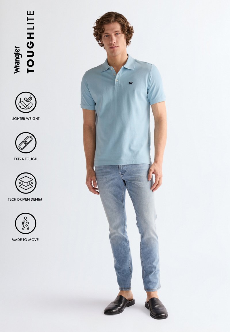 Muž oblečený v svetlomodrej polokošeli, svetlo vybledlých rifliach a čiernych slip-on topánkach, stojaci na jednotnom bielom pozadí.