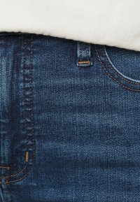 Mörkblå denimjeans med orange sömmar, en framficka och en något texturerad yta. Midjan täcks delvis av en vit skjorta.