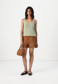 Lys grønn strikket tanktop, brune semskede shorts, svarte leiligheter med nagler, og en liten teksturert brun veske. Modellen står mot en hvit bakgrunn.