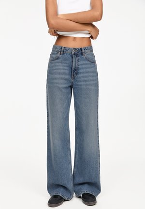 Wijduitlopende blauwe denim jeans met een hoge taille, voorkeurszakken en subtiele vervaging, gecombineerd met zwarte schoenen en een aansluitend wit topje.