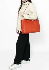Borsa a tracolla arancione in pelle con un design strutturato, caratterizzata da una base piatta, due manici superiori e un'etichetta rimovibile. Texture liscia e minimalismo nei dettagli hardware.
